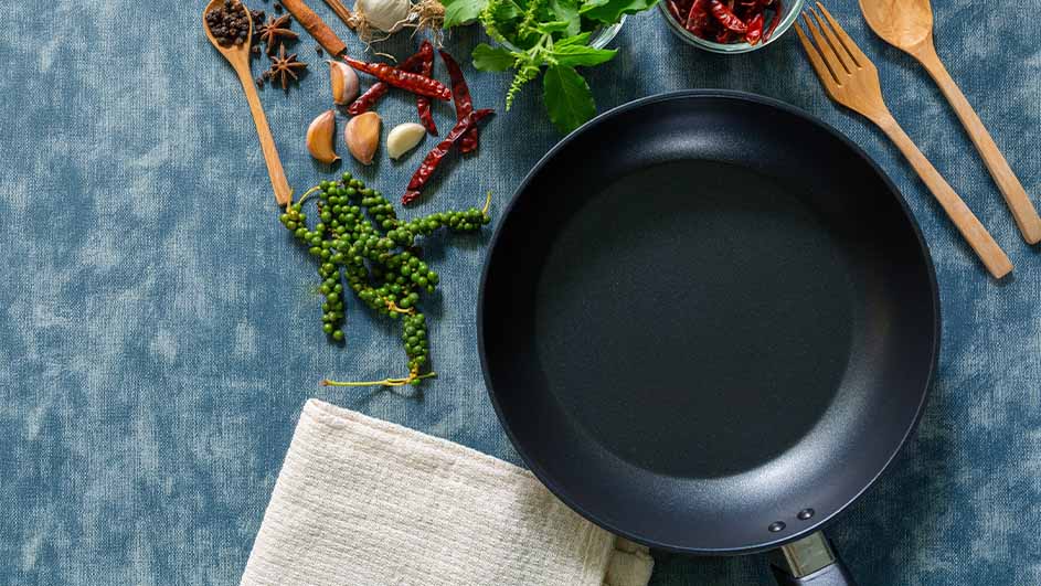 Een zwarte koekenpan ligt op een blauwe ondergrond met kruiden, knoflook, pepers en houten keukengerei.