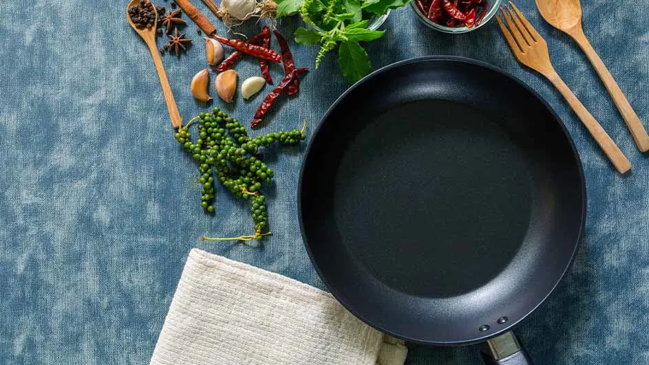 Een zwarte koekenpan ligt op een blauwe ondergrond met kruiden, knoflook, pepers en houten keukengerei.