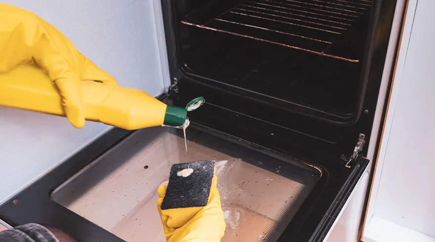 Gele handschoenen houden een spons vast in een oven, reinigingsmiddel wordt aangebracht. De oven is zwart en de binnenkant is vies.