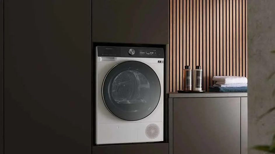 Witte wasdroger met donkere deur in een donkere kast. Naast de droger staan handdoeken en twee flessen op een plank.