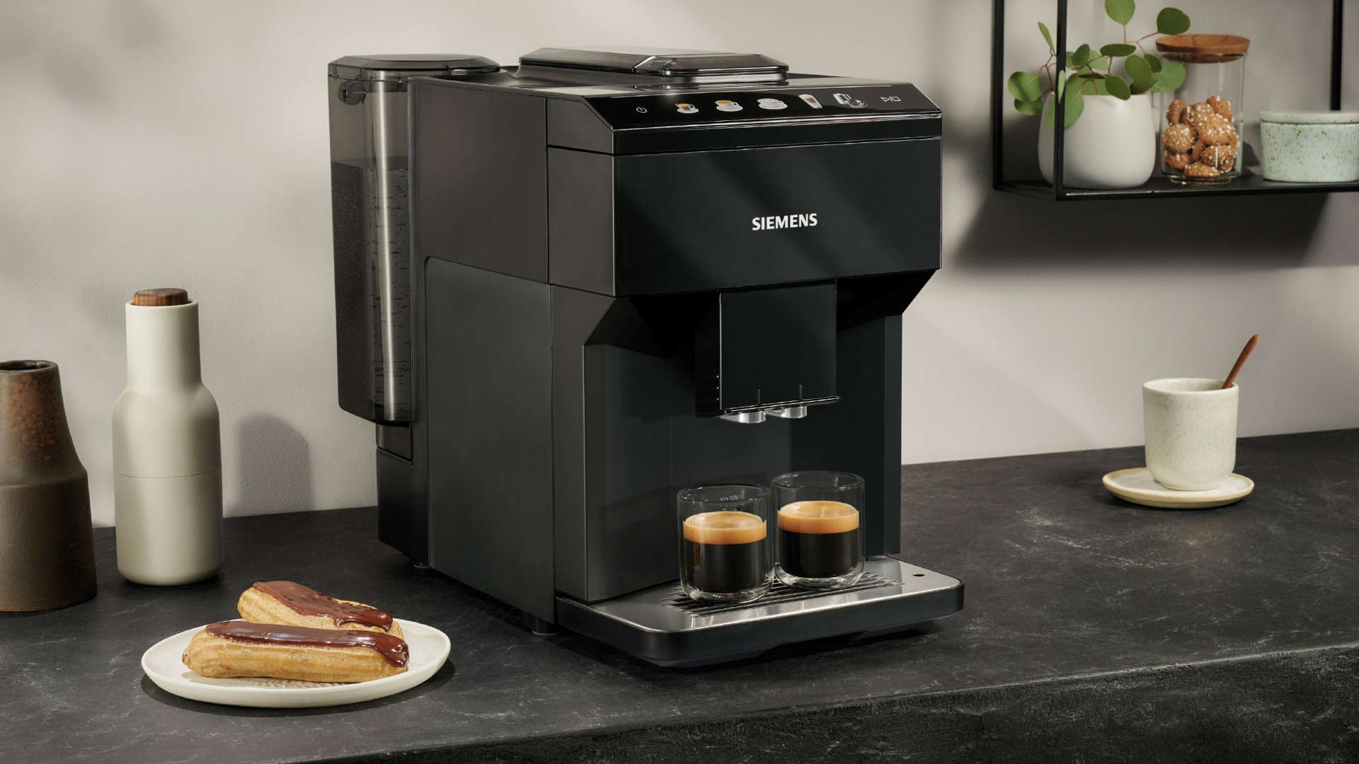 Zwarte Siemens koffiemachine met twee glazen espresso. Op de voorgrond een bord eclairs. Rek met potten op de achtergrond.