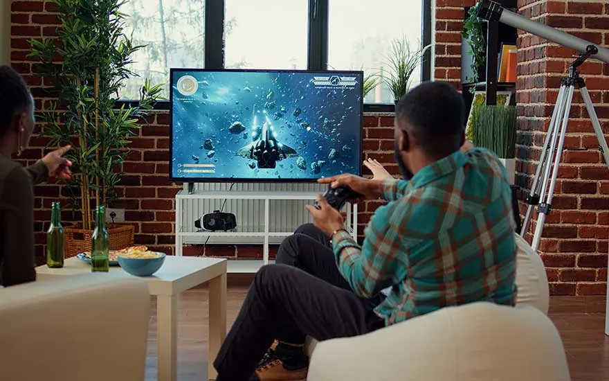 Een man en een vrouw gamen samen op een grote tv in een moderne woonkamer, omringd door planten, snacks en spelaccessoires.