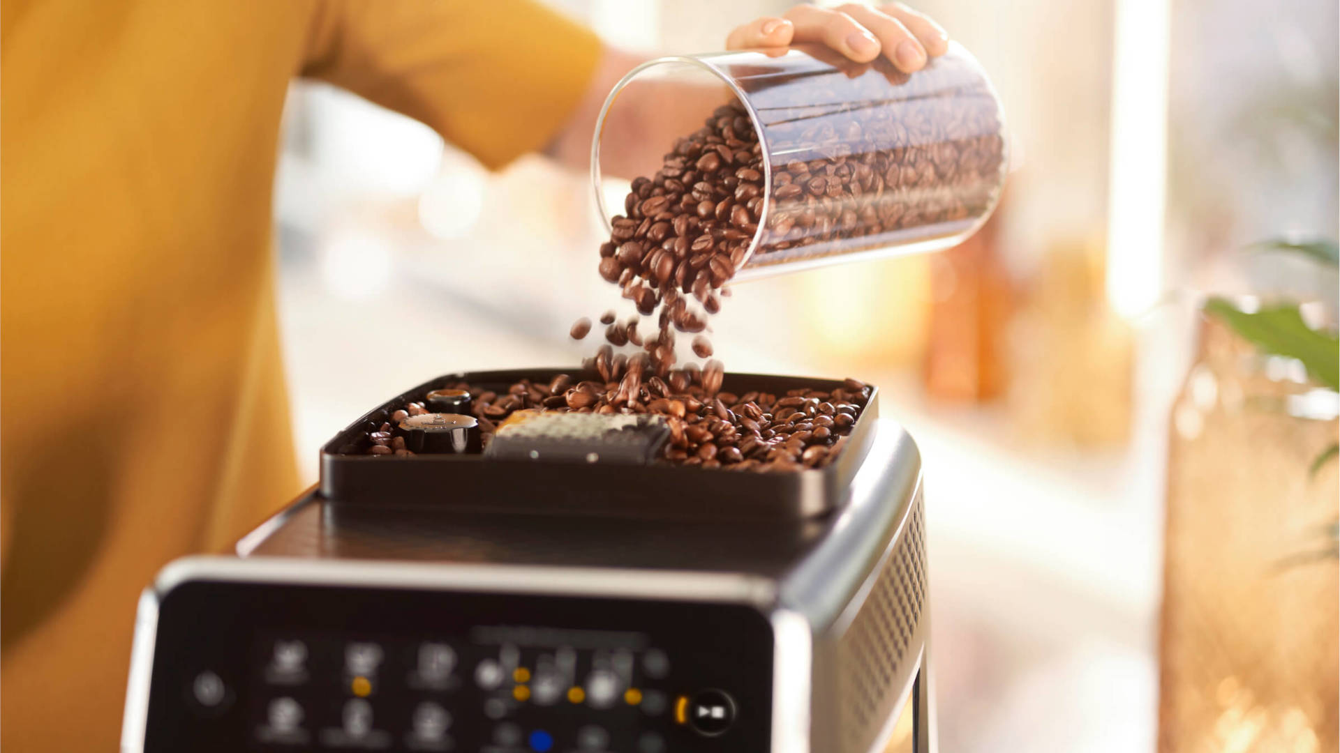 Een persoon giet koffiebonen uit een glazen pot in een zwarte koffiemachine. Op de achtergrond is een kamer met een plant te zien.