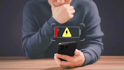 Een persoon houdt een zwarte telefoon vast onder een batterijpictogram met een rode balk en een gele bliksemschicht.