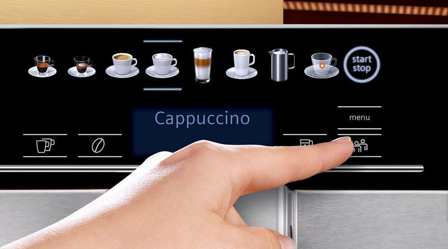 Hand drukt op knop 'personen' op een koffiezetapparaat met zwarte display en pictogrammen van dranken. 'Cappuccino' staat op scherm.