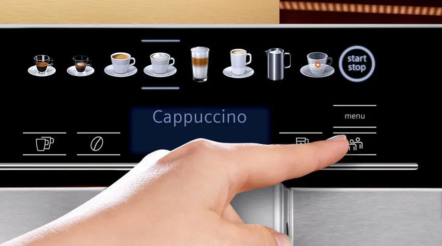 Hand drukt op knop 'personen' op een koffiezetapparaat met zwarte display en pictogrammen van dranken. 'Cappuccino' staat op scherm.