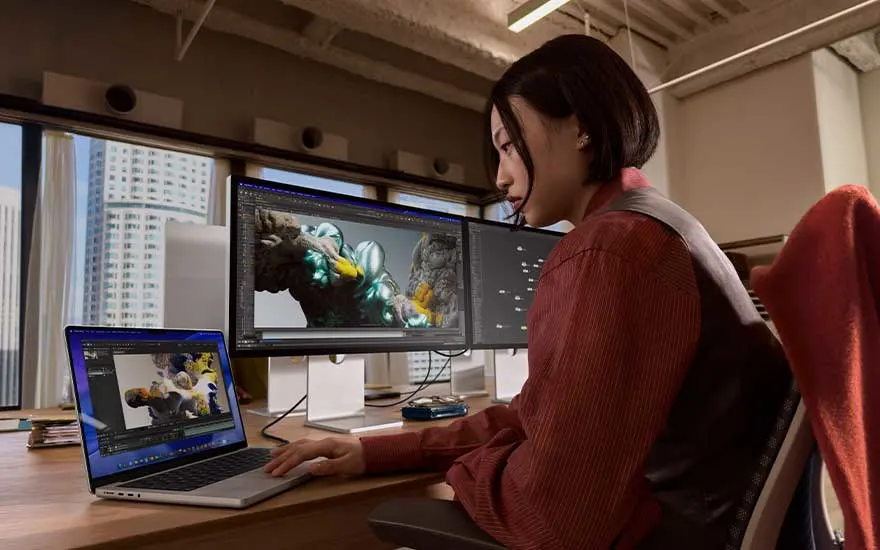 Persoon zit aan een bureau in een modern kantoor, werkt geconcentreerd op een MacBook verbonden met groot scherm, waarop 3D animaties worden bewerkt.