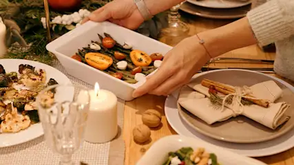 Gedekte tafel met een wit gerecht met asperges, tomaten en mozzarella. Handen tillen het gerecht op. Kaarsen en gerechten op tafel.