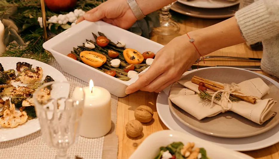Gedekte tafel met een wit gerecht met asperges, tomaten en mozzarella. Handen tillen het gerecht op. Kaarsen en gerechten op tafel.