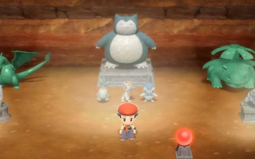 In een ondergrondse kamer staat een kleine trainer tussen grote Pokémon beelden, met een reusachtige Snorlax sculptuur centraal op een stenen sokkel.