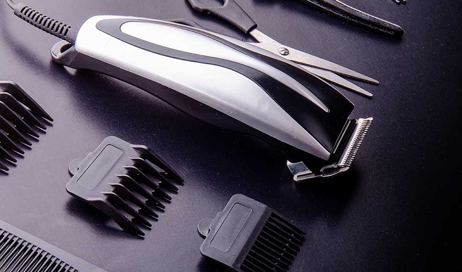 A hair clipper lies on a dark surface among guide combs, scissors, and other barber accessories.