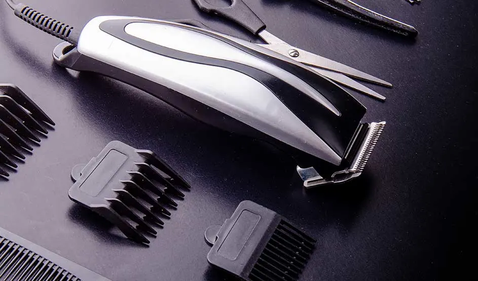 A hair clipper lies on a dark surface among guide combs, scissors, and other barber accessories.