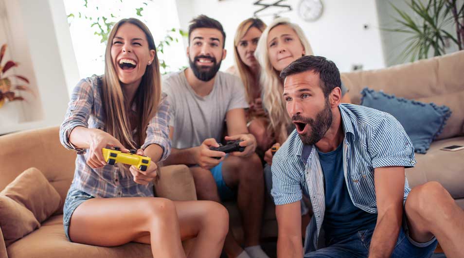 Een groep vrienden zit op een bank en geniet enthousiast van een videogame, allemaal lachend en geconcentreerd op de actie.