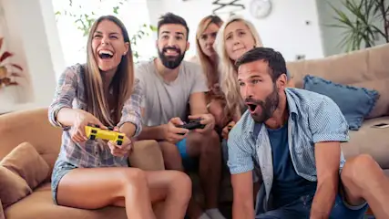 Een groep vrienden zit op een bank en geniet enthousiast van een videogame, allemaal lachend en geconcentreerd op de actie.