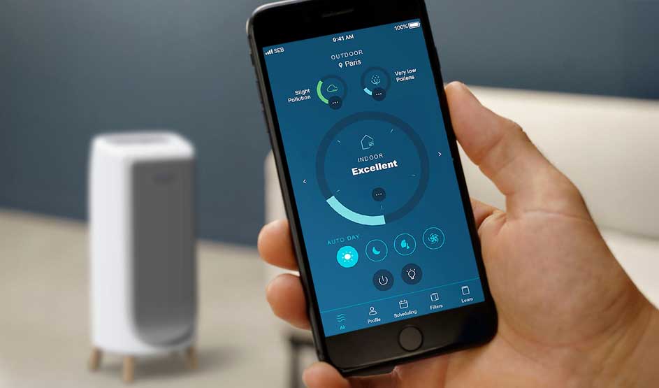 Hand holding smartphone displaying air quality monitoring app with excellent indoor rating, white air purifier in background