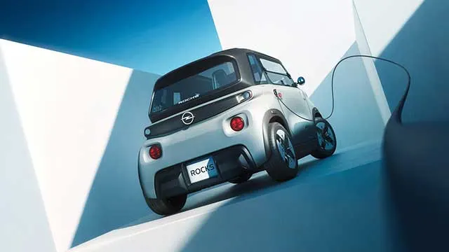 Achteraanzicht van de compacte elektrische Opel Rocks die aan het opladen is, in een lichtblauwe architecturale setting met sterke schaduwen.