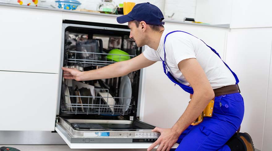 Een monteur in blauwe overall en pet knielt voor een open vaatwasser in een witte keuken.