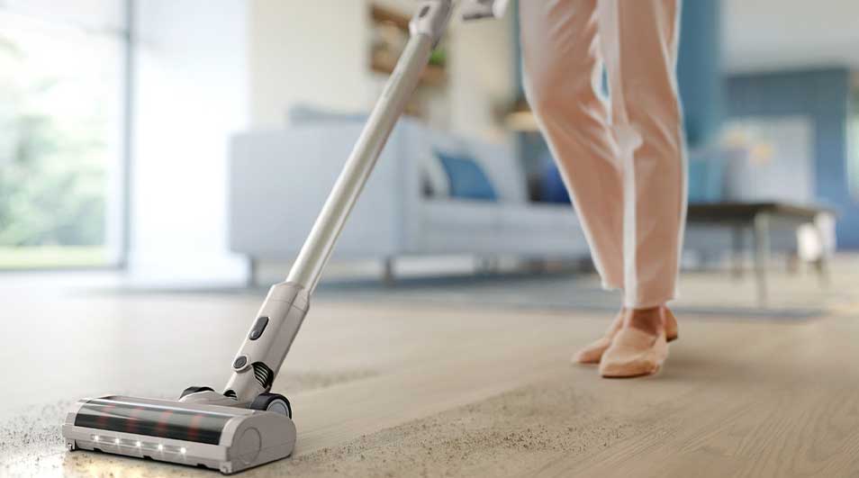 Iemand stofzuigt een houten vloer met een lichte steelstofzuiger, waarbij een strook kruimels netjes wordt opgezogen in de zonnige woonkamer.