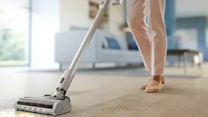 Iemand stofzuigt een houten vloer met een lichte steelstofzuiger, waarbij een strook kruimels netjes wordt opgezogen in de zonnige woonkamer.