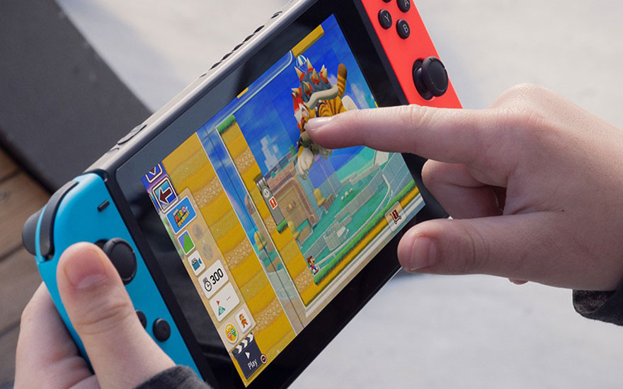 Handen die een Nintendo Switch vasthouden met blauwe en rode Joy-Cons, spelend een kleurrijke game op het touchscreen in handheld-modus.