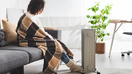 Vrouw zit op grijze bank met plaid, bij kachel en plant. Bureau met stoel staat verderop in de kamer.