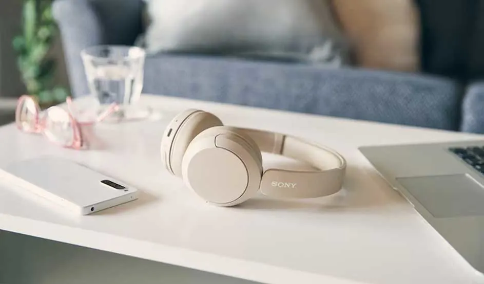Op een witte tafel liggen beige Sony koptelefoon, een smartphone, roze bril en een laptop, met een glas water en een blauwe bank op de achtergrond.