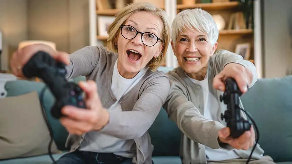 Twee oudere vrouwen zitten op een bank en gamen enthousiast met gamecontrollers in hun handen. Ze lachen breed en lijken veel plezier te hebben.