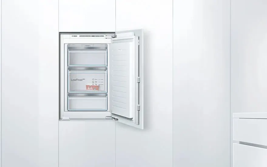White built-in freezer in a sleek kitchen wall, door open so that three transparent drawers with LowFrost label and food packaging are visible.