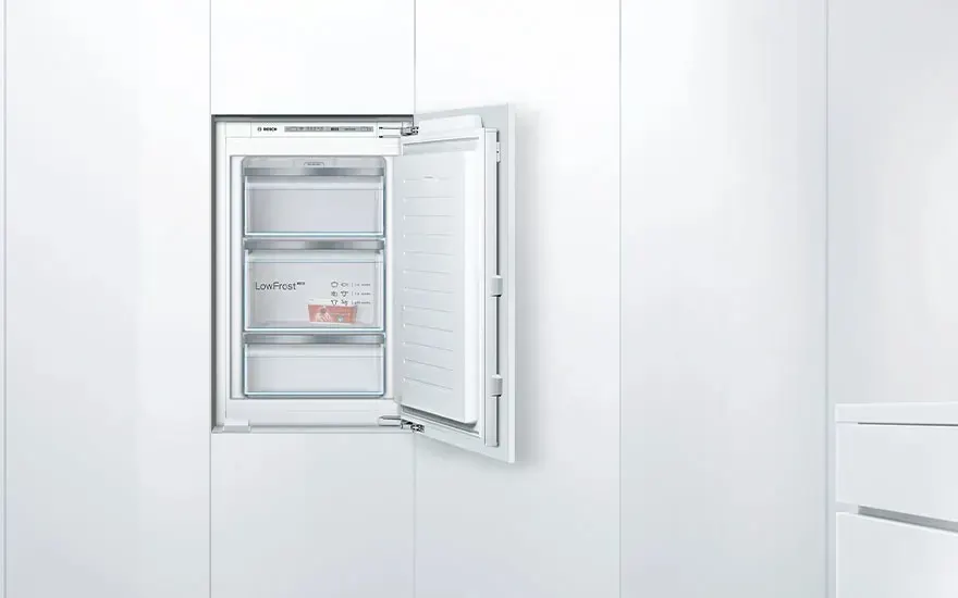 White built-in freezer in a sleek kitchen wall, door open so that three transparent drawers with LowFrost label and food packaging are visible.