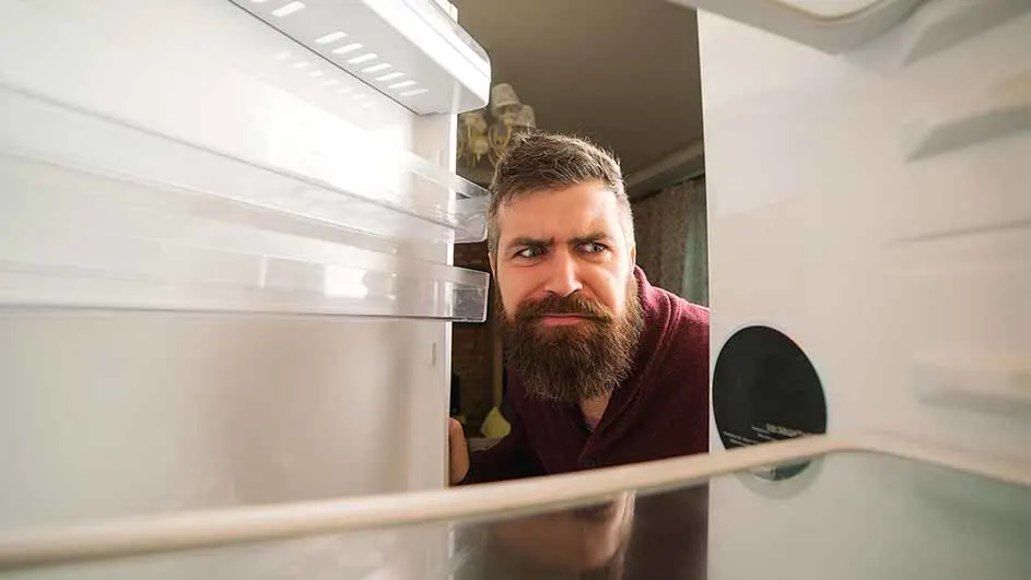 Een man met een baard kijkt in een lege, witte koelkast. De man draagt een bordeauxrood shirt.
