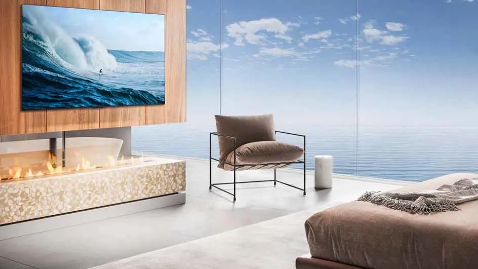 Moderne woonkamer met een aan de muur gemonteerde tv waarop een surfer op een golf te zien is, naast een haard en een groot raam met uitzicht op de oceaan.