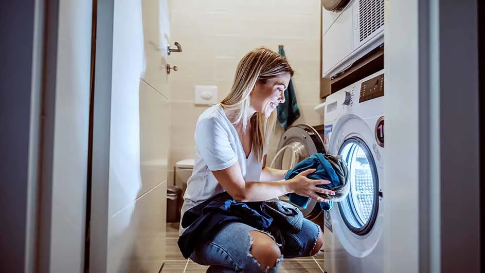 Een vrouw hurkt bij een wasmachine en haalt blauwe kleding eruit in een badkamer.