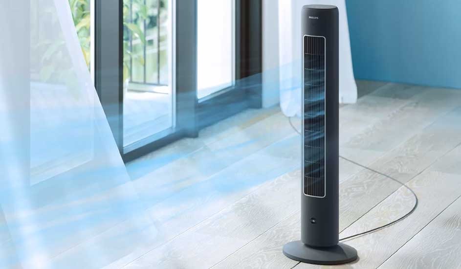 Slim black tower fan blows cool air in a bright room with wooden floor and open window with flowing curtain.