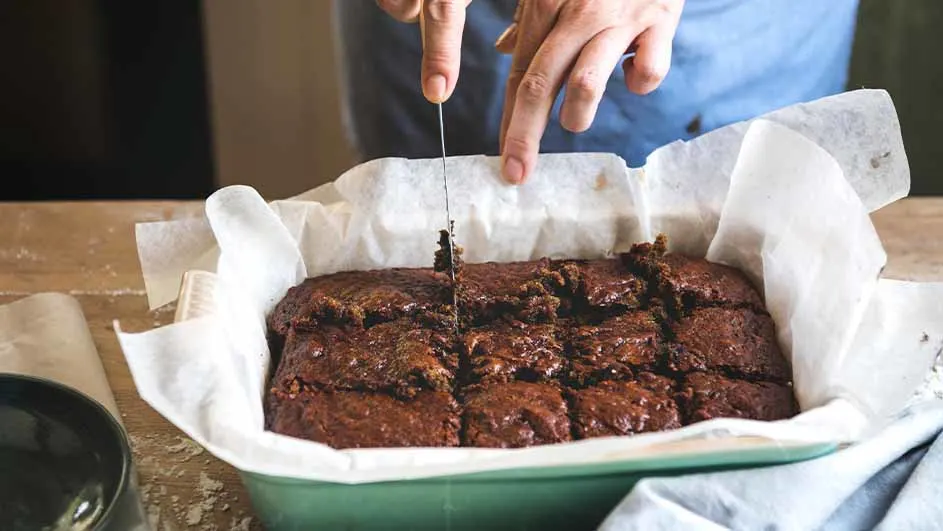 Een persoon snijdt brownies in een groene bakvorm bekleed met bakpapier met een mes.