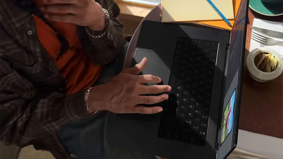 Man gebruikt laptop, met notities, bestek en zoutvaatje op tafel. Hij draagt een geruit overhemd over een oranje shirt.