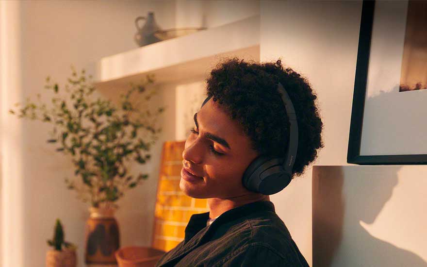 Jongeman met draadloze koptelefoon leunt glimlachend achterover in warme zon, in een gezellig interieur met planten.