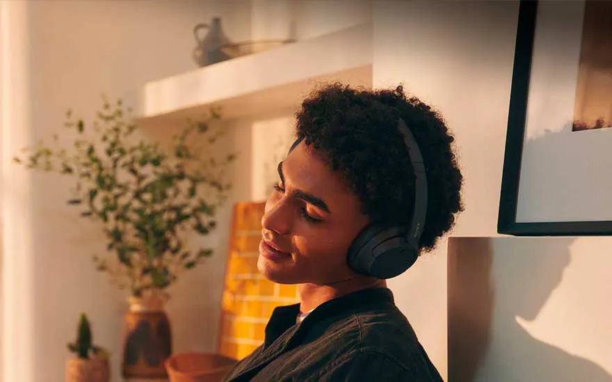 Jongeman met draadloze koptelefoon leunt glimlachend achterover in warme zon, in een gezellig interieur met planten.