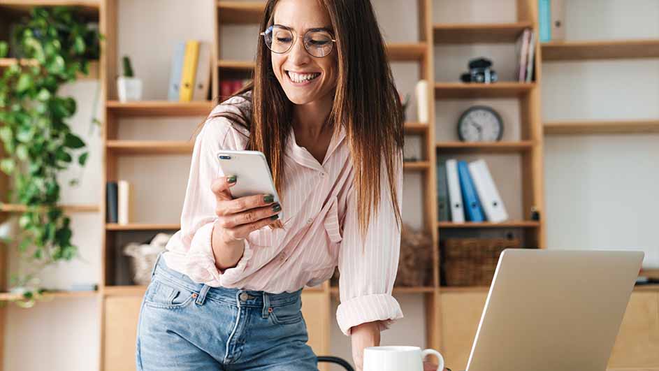 Vrouw met bril en roze blouse kijkt naar een witte smartphone bij een laptop en een witte mok. Op de achtergrond een houten boekenkast.