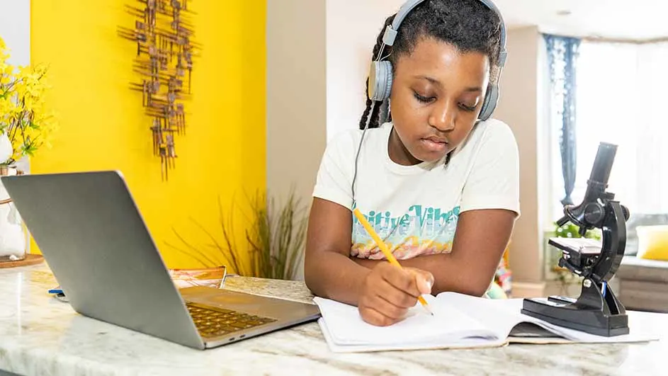 Een persoon met een koptelefoon zit aan een tafel met een laptop, een microscoop, een notitieboek en een potlood.