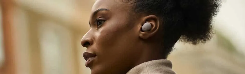 A person is wearing a beige Samsung wireless earbud in their right ear. The background appears outdoors and softly blurred.