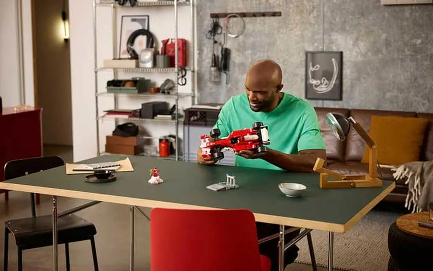 Man in groene trui zit aan een moderne tafel en bekijkt een rode LEGO raceauto, in een stijlvolle woonkamer met lamp.