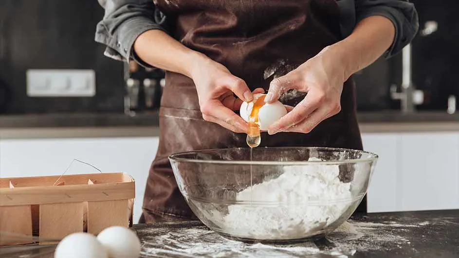 Iemand breekt een ei in een glazen kom met bloem, draagt een bruin schort en staat aan een tafel.