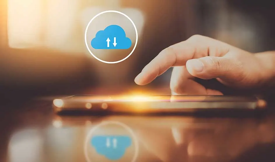 Een hand raakt een smartphone aan op een tafel, met een blauwe cloud upload/download icoon erboven. Geen merk zichtbaar.