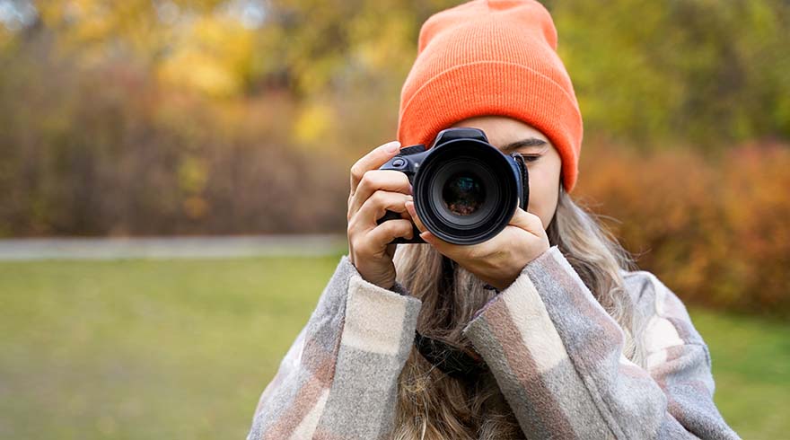 Een persoon met een oranje muts houdt een camera vast in een park met bomen op de achtergrond.