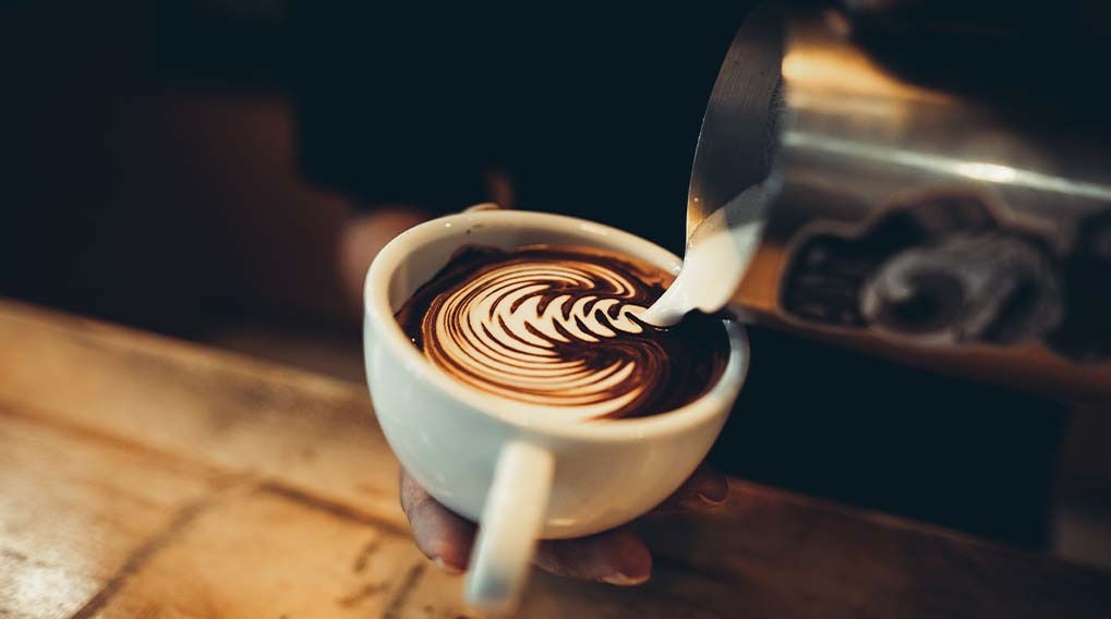 A white cup on a wood surface holds coffee with a swirling design created by a silver pitcher pouring in white cream.
