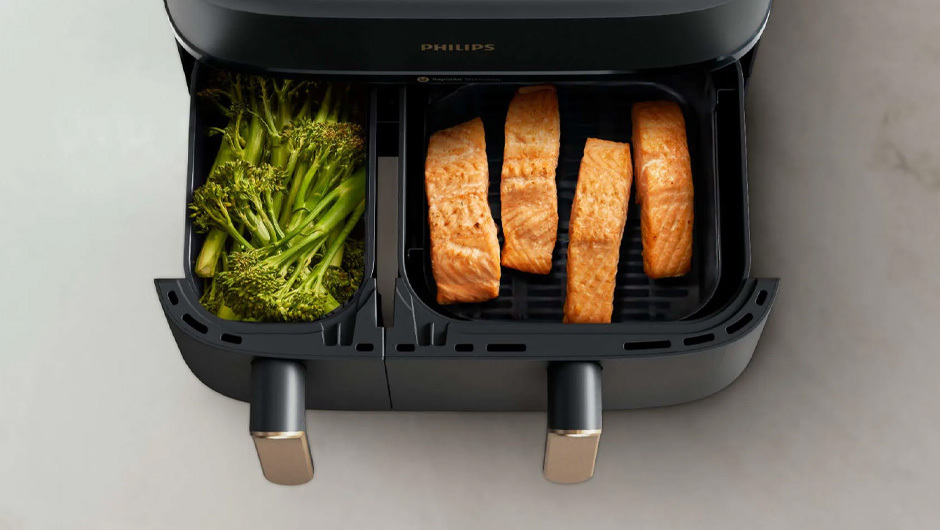 Een zwarte airfryer met twee compartimenten, één met groene broccoli en de andere met vier stukken oranje zalm.