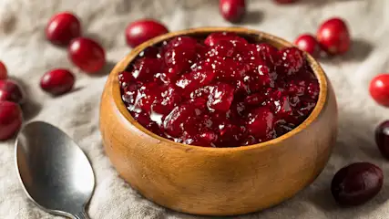 Een houten kom vol rode cranberrysaus staat naast een lepel op een grijze doek, met losse rode cranberries eromheen.