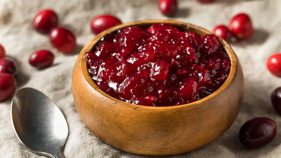 Een houten kom gevuld met rode cranberrysaus staat naast losse cranberries en een zilveren lepel op een grijze linnen ondergrond.