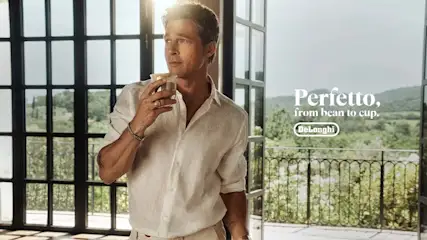 Man in wit linnen overhemd houdt een beker vast, met een landschap en het DeLonghi-logo op de achtergrond.