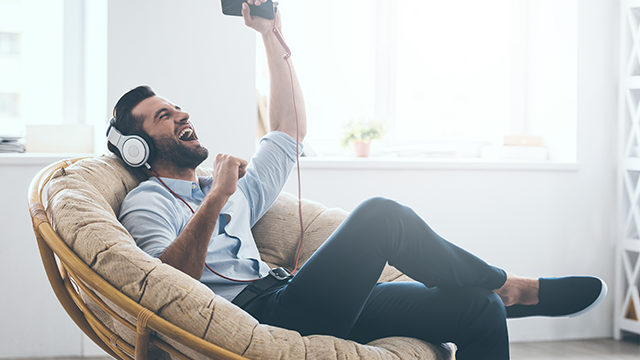 Een man zit in een stoel met een koptelefoon op, zijn arm omhoog met een telefoon in zijn hand.